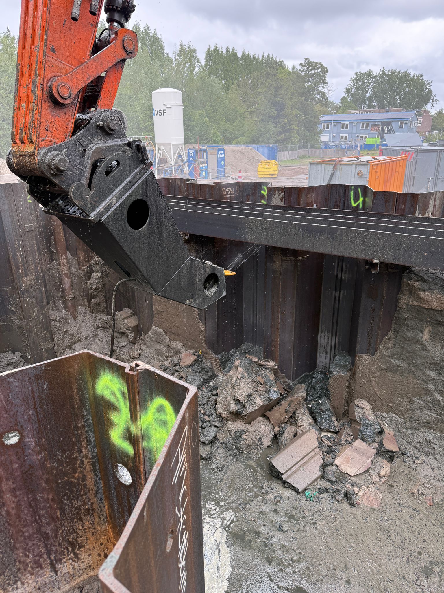 Hompert-Renes - Damwand reinigen | Hompert - Renes excavator bucket gripping rusted steel sheet piles at a construction site with dirt and rubble nearby.