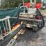 A compact drill rig on tracks with hoses and metal framing at a construction site, on muddy ground with wooden planks nearby.