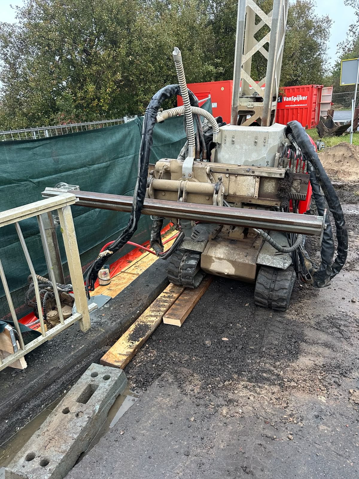Hompert-Renes - Schampkant Emmeloord | Hompert - Renes A compact drill rig on tracks with hoses and metal framing at a construction site, on muddy ground with wooden planks nearby.